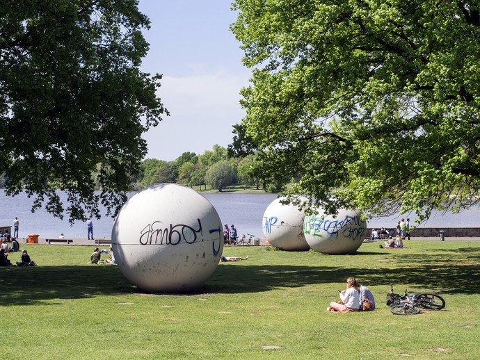 You see giant pool balls in a parc with trees and a lake in the background. You see giant pool balls in a parc with trees and a lake in the background.